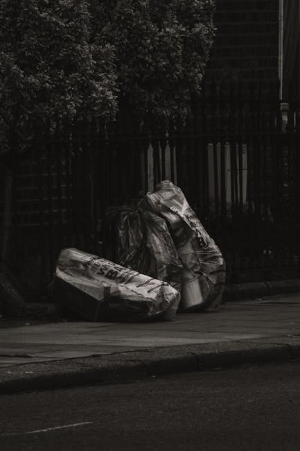 A close-up view of a person wearing a white work glove and a silver wristwatch, using a rusty shovel to lift a bundle of garden and outdoor debris into a large white polypropylene sack. The debris con