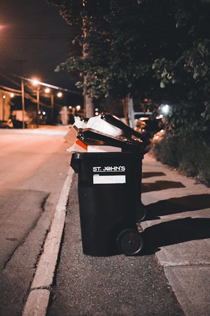 A person disposing of paper waste inside a white recycling bin with a black label, using their right hand to place a crumpled brown paper bag into the bin, which is positioned on a wooden surface. In 