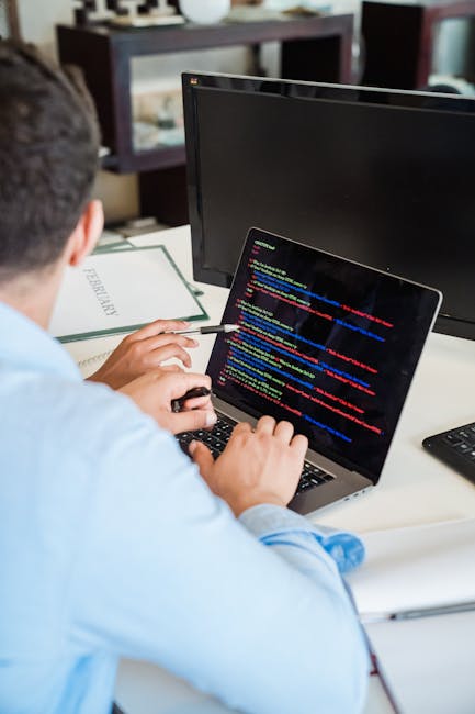 A person seated at a desk is working on a laptop computer with a black screen displaying lines of multicolored programming code. The individual, wearing a light blue shirt, is holding a stylus or pen 